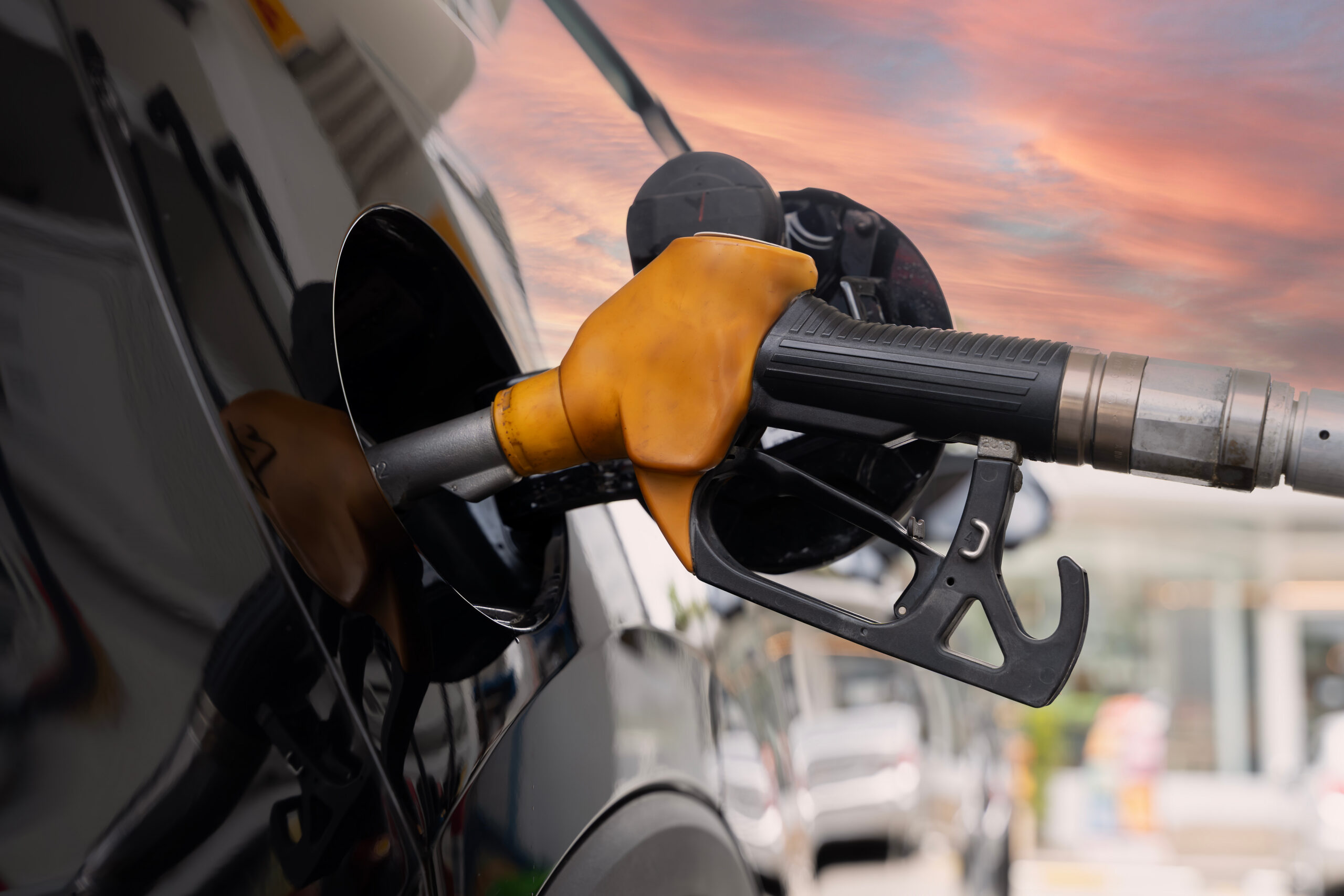 fuel nozzle refueling black car at gas station with sunset sky reflection on surface, close up detail